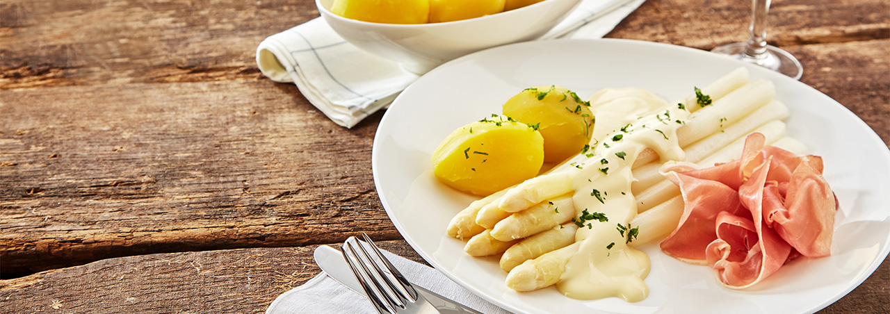 Spargel mit Sauce Hollandaise, dazu Kartoffeln und Schinken. Daneben ein Glas Weißwein