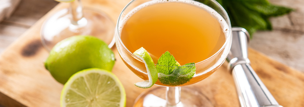 Old Cuban-Cocktail auf Holztisch, daneben eine halbierte Zitrone, im Hintergrund Minze