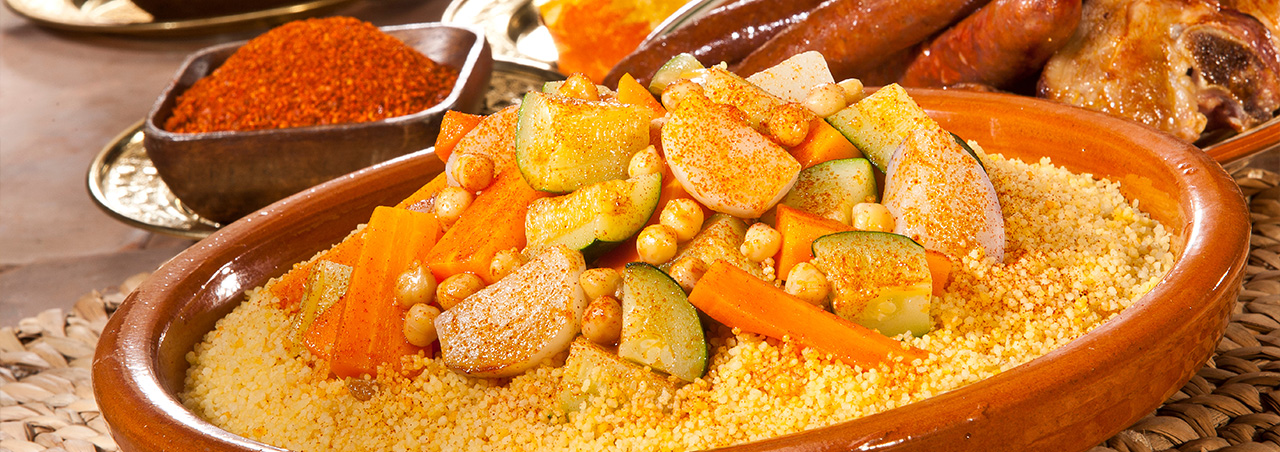 Nahaufnahme von Merguez-Couscous in traditionellem Berber-Teller, im Hintergrund verschiedene Couscous-Gewürze 