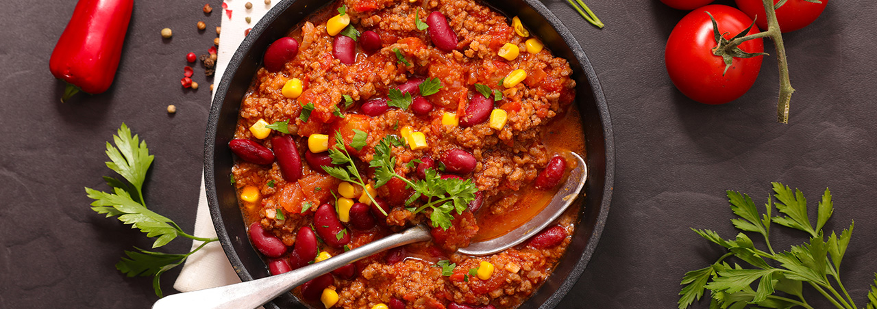 Chili con carne mit Löffel in rundem Teller auf schwarzem Untergrund, im Hintergrund Tomaten