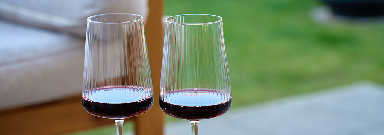 Zwei stilvolle Rotwein-Gläser auf gepflegtem Holztisch, im Hintergrund Gartenmöbel und grüner Rasen
