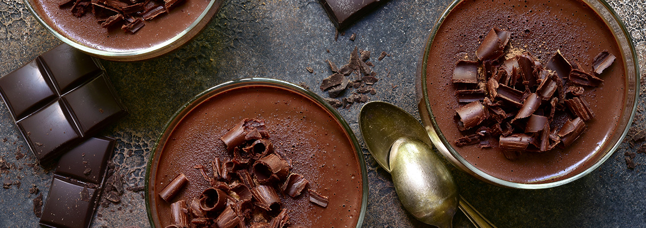 Edel angerichtete Mousse au Chocolat, daneben schwarze Schokoladen-Carrés und goldene Dessert-Löffel
