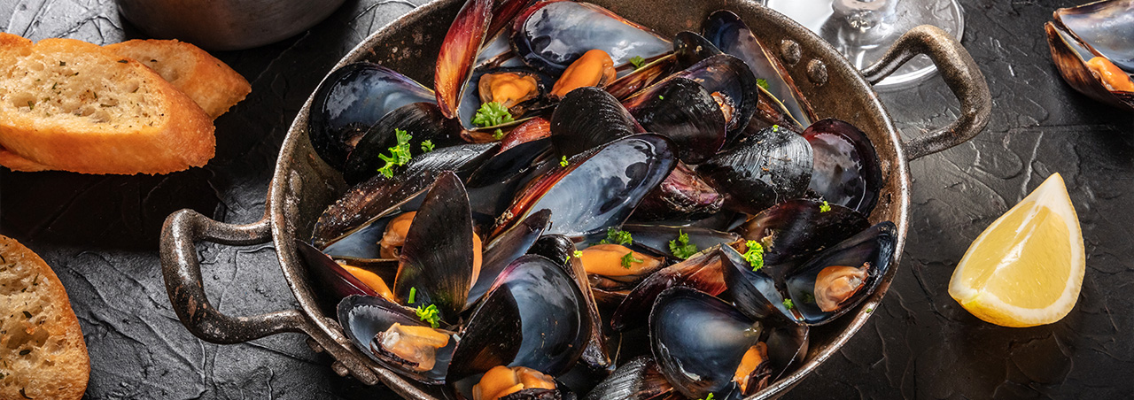 Moules marinières in Edelstahltopf auf schwarzem Tisch. Dazu heller Roséwein, Pommes und gespaltene Zitronen