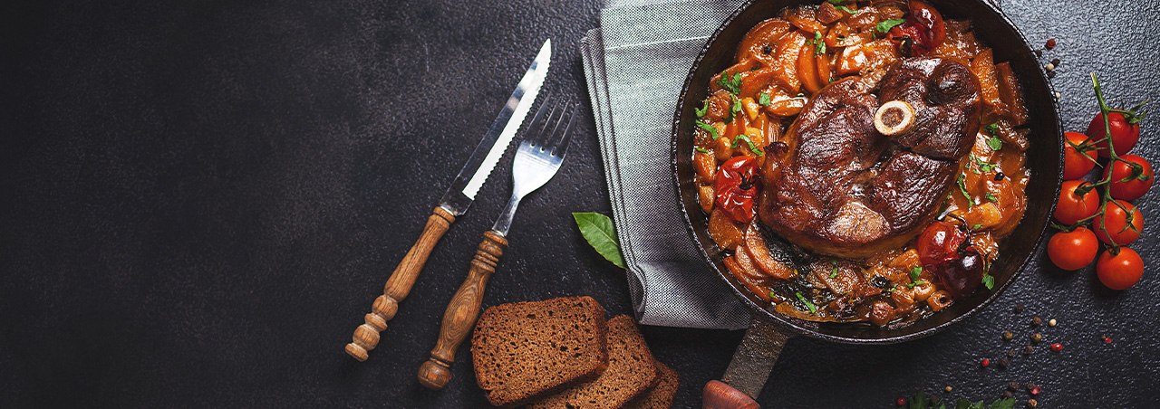 Osso Bucco in Eisengusspfanne auf schwarzer Platte, daneben traditionelles Besteck, Kirschtomaten und Krätuer