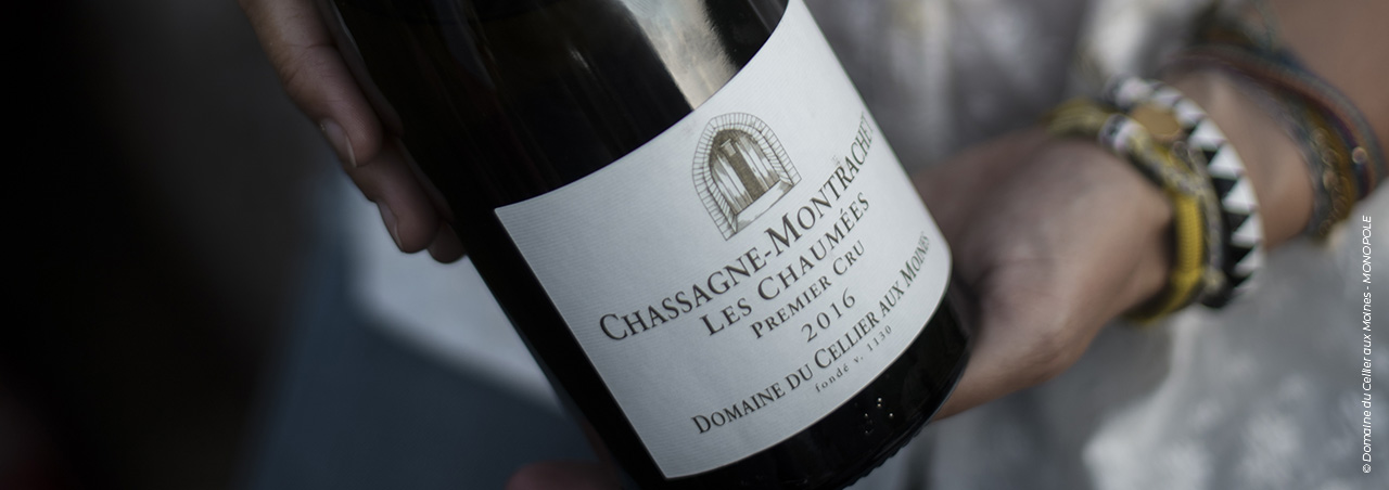 Eine Aufnahme von seltener Eleganz, auf der zwei Hände behutsam eine Flasche Chassagne-Montrachet des Domaine du Cellier aux Moines halten.