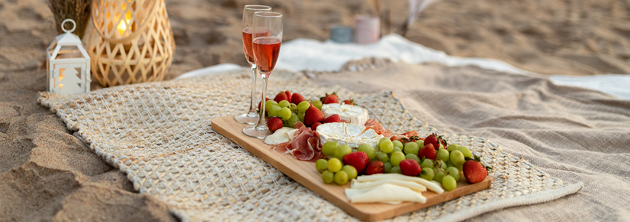 Romantisches Picknick mit zwei Gläsern Rosé-Champagner, frischen Früchten und verschiedenen Käsesorten am Strand.