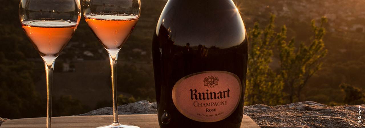 © Ruinart, eine Flasche Ruinart Rosé-Champagner mit zwei gefüllten Champagnergläsern vor stimmungsvollem Sonnenuntergang