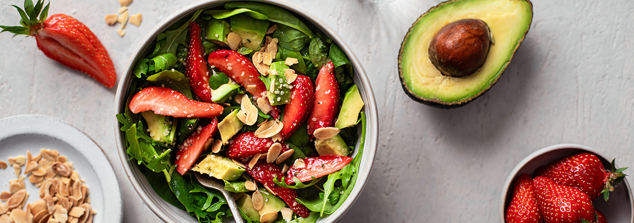 Avocado-Erdbeersalat in runder Schale auf dezent dekoriertem Untergrund, daneben ein Glas Wasser 
