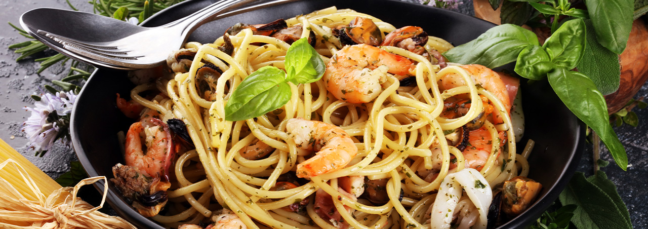Mediterrane Meeresfrüchte-Pasta, angereichert mit Basilikum-Blättern. Serviert in schwarzem Teller auf schwarzer Schiefernplatte.