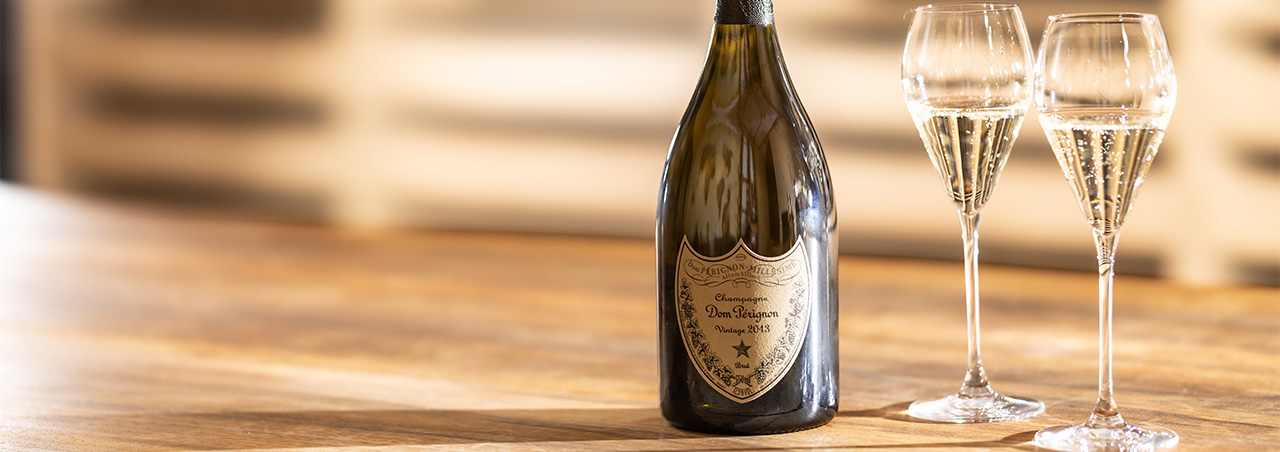Eine Flasche Dom Pérignon Vintage 2013, daneben zwei gefüllte Champagnergläser auf Holztresen