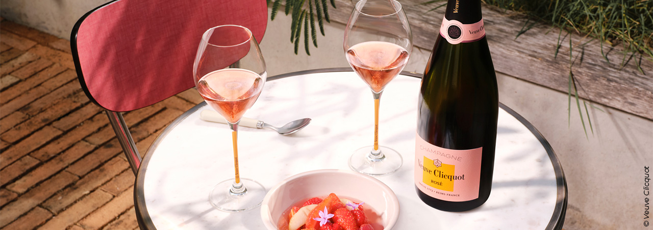 © Veuve Clicquot, Brut Rosé-Champagnerflasche von Veuve Clicquot mit zwei gefüllten Champagnergläsern auf gepflegt gedecktem Tisch, dazu Erdbeeren in einer Schale