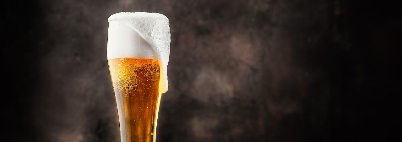 Ein Glas frisches Bier auf Holztisch vor dunklem Hintergrund. Der Schaum quillt genüsslich über
