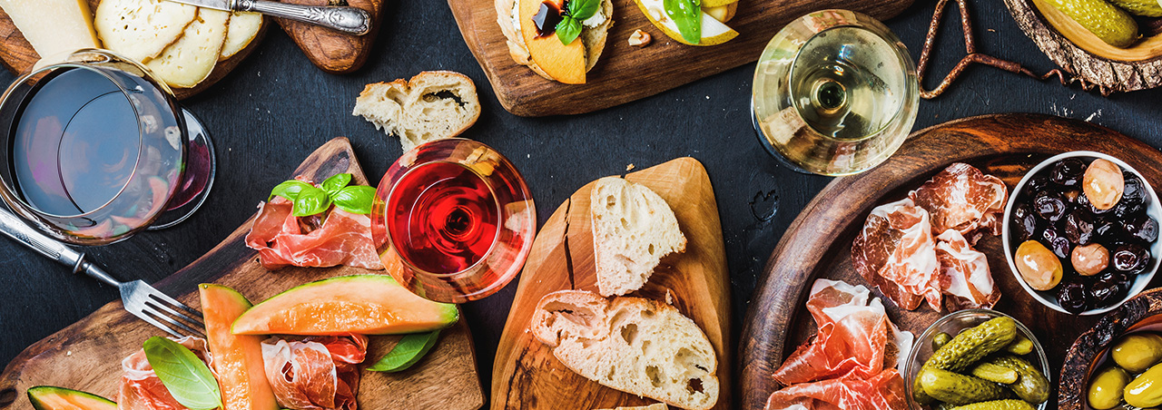 Italienisches Antipasti-Wein-Snack-Set. Bruschetta, verschiedene Käsesorten, mediterrane Oliven, eingelegtes Gemüse, Prosciutto di Parma mit Melone, Salami und Wein in Gläsern vor einem schwarzen Hintergrund