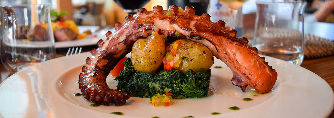 Gegrillter Oktopus mit Kartoffeln, dahinter ein gefülltes Rotweinglas