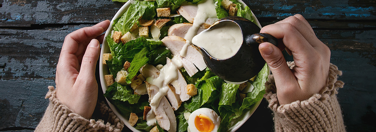 Runde Schale mit Caesar Salad auf anthrazitfarbenem Holztisch; Frau gießt helle Sauce darüber; welcher Wein passt hier?