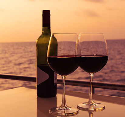 Zwei gefüllte Rotweingläser, daneben eine Flasche Rotwein; im Hintergrund ein Sonnenuntergang über dem Meer