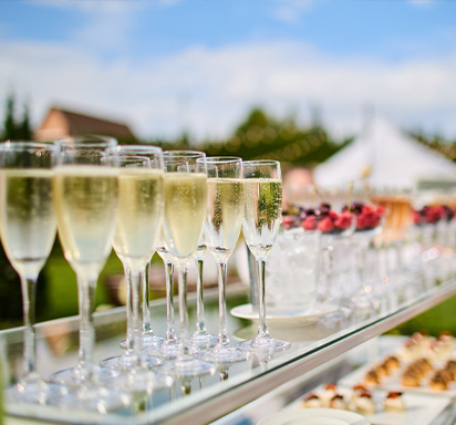 Reihen von Sekt- oder Champagnergläsern, gefüllt mit Schaumwein, auf einem gläsernen Servierwagen bei einem Hochzeitsempfang im Freien