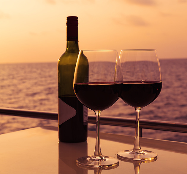Zwei gefüllte Rotweingläser, daneben eine Flasche Rotwein; im Hintergrund ein Sonnenuntergang über dem Meer